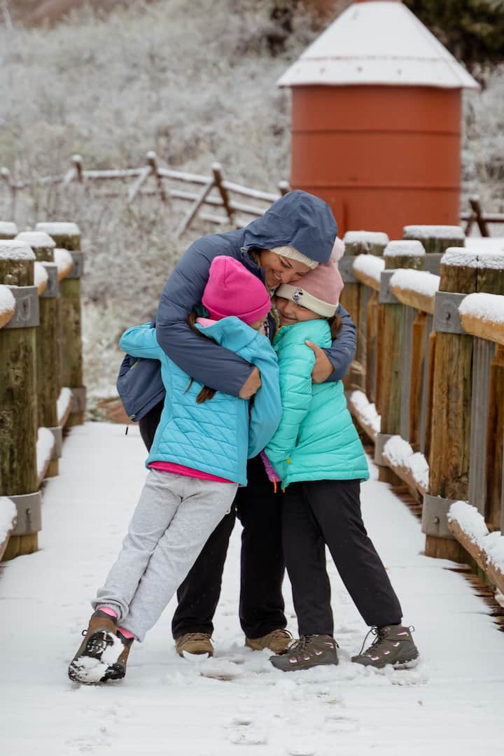 Two children in bright winter jackets and hats embrace an adult wearing a hooded jacket on a snow-covered wooden bridge, with frosty trees and a red silo visible in the background.