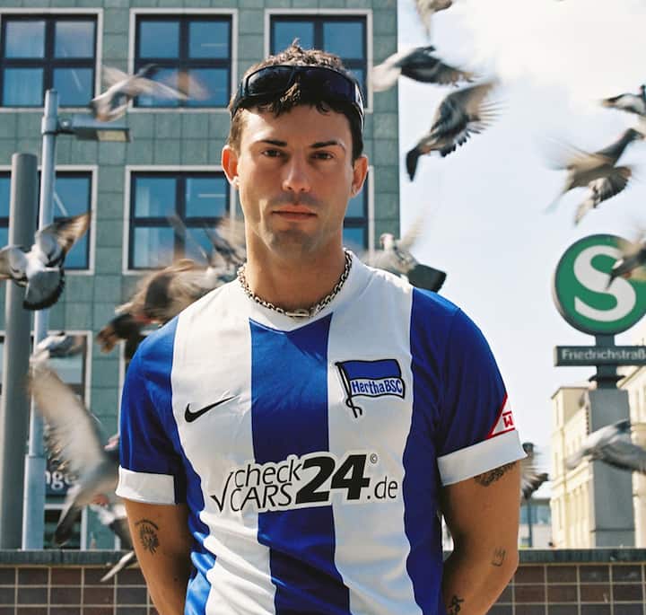 Person wearing a Hertha BSC soccer jersey with blue and white stripes, surrounded by pigeons in motion, with a cityscape and a green S-Bahn sign reading 'Friedrichstraße' in the background.