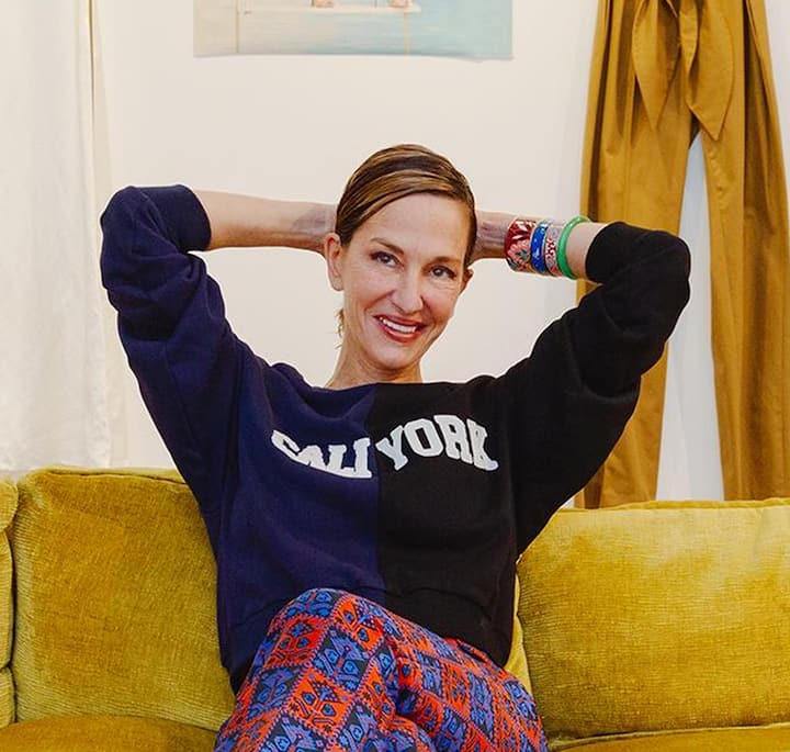 Person reclining on a mustard yellow couch, wearing a black and navy sweatshirt with split text that reads 'CALI YORK' and colorful patterned pants. Bright bracelets adorn their wrist, and a relaxed atmosphere is suggested by the curtains and wall art in the background.