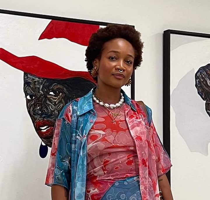 Art gallery setting showing a person dressed in red and blue patterned clothing, with two framed paintings behind them—one featuring a stylized face with a red hat and bold colors.