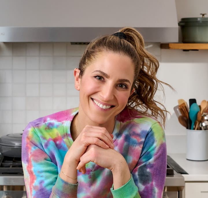 A kitchen setting featuring a person in a tie-dye sweatshirt with their hands clasped, surrounded by a stovetop, cookware, and a container of utensils on the counter.