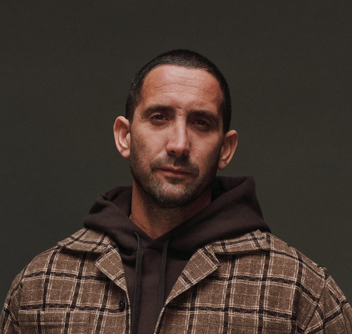 Individual dressed in a checkered brown jacket layered over a dark hoodie, set against a plain dark backdrop.