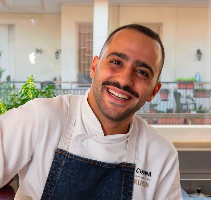 A chef wearing a white uniform and denim apron with 'CUINA' and 'RUBEN' embroidered on it, standing in a bright kitchen with plants and colorful pots visible in the background.