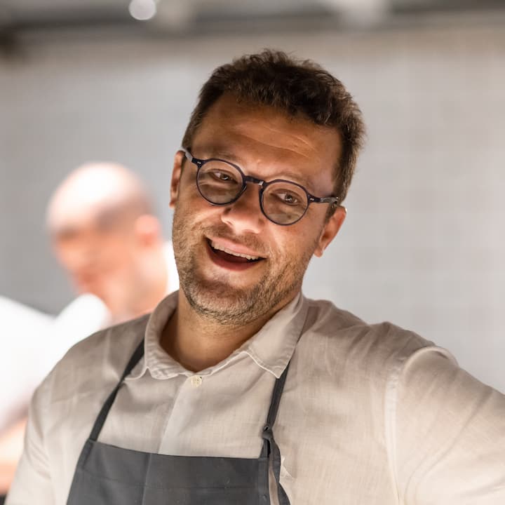 Close-up of a person dressed in a white shirt and black apron in a professional kitchen setting, with tiled walls visible in the background.