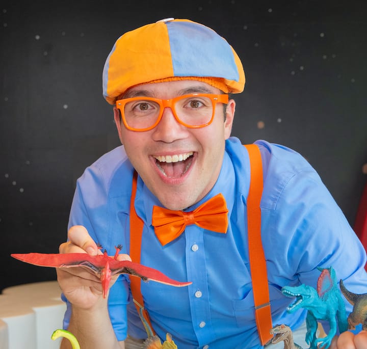 A person dressed in a bright outfit with an orange and blue hat, orange suspenders, and an orange bow tie, holding a red pterodactyl toy among other colorful dinosaur figurines on a table.