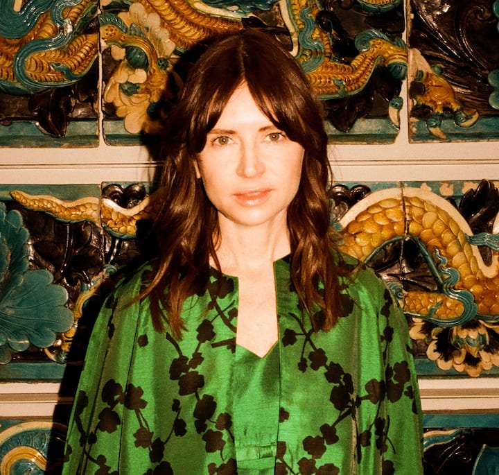 A person wearing a vibrant green floral-patterned outfit stands in front of an ornate wall featuring colorful dragon motifs and intricate tile designs.