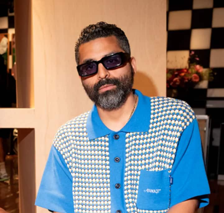 Close-up of a person wearing a retro-style blue and beige patterned shirt, with a modern interior featuring flowers and checkered decor in the background.