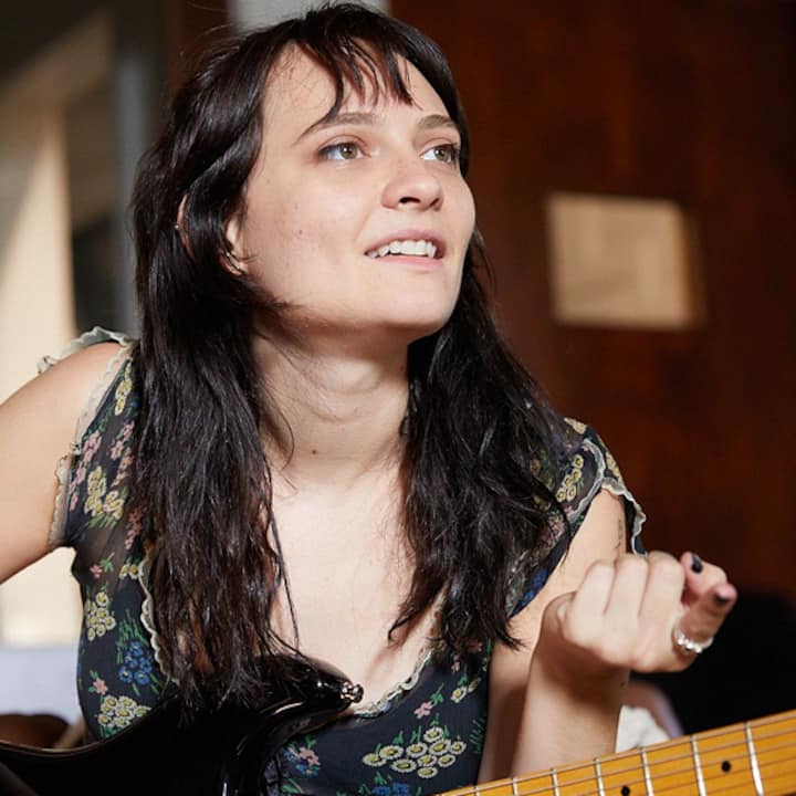 A person with long dark hair strumming a black electric guitar, seated indoors with a blurred background featuring warm lighting and wooden textures.
