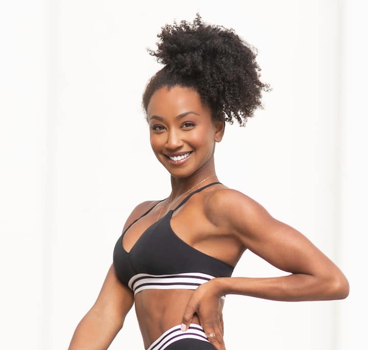 A fit woman with curly hair wearing a black sports bra and matching striped athletic leggings, posing confidently against a bright white background.