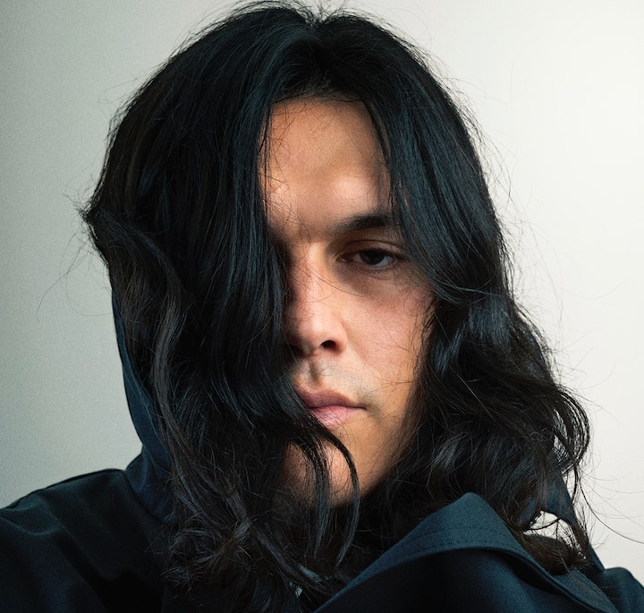 Close-up of dark wavy hair cascading over a black hooded garment, set against a light background.