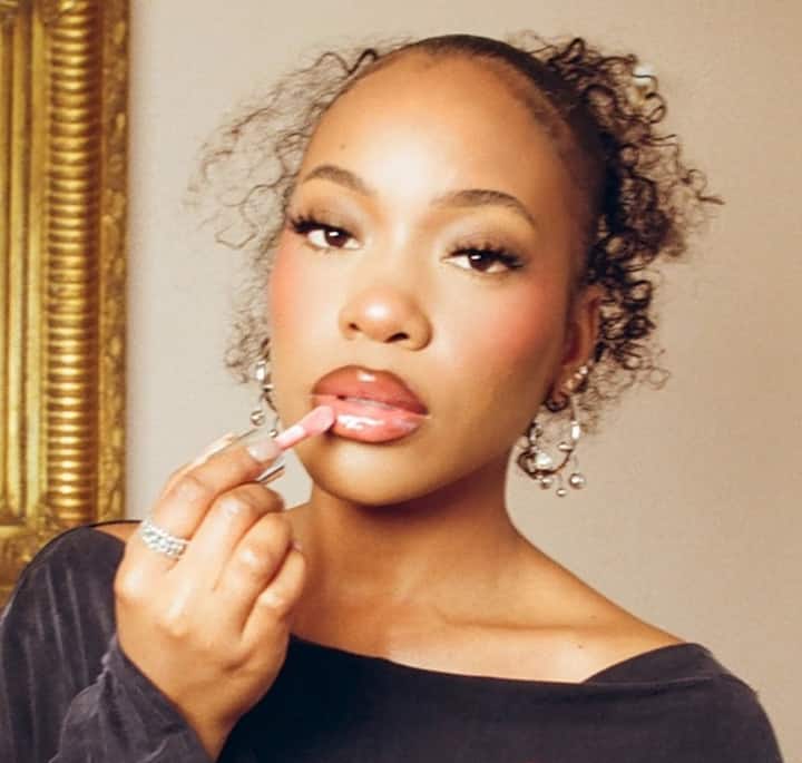 Close-up of a person wearing a black top and silver rings, holding a small item near their face, with a gold-framed decorative mirror visible in the background.