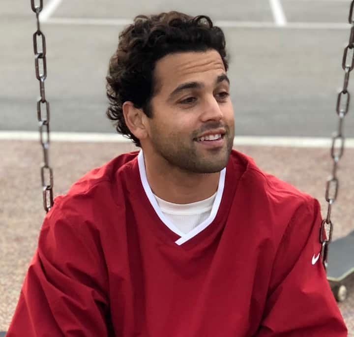 A person in a red sweater with white trim and Nike branding sits on a swing in a park, with chains and background pavement visible.