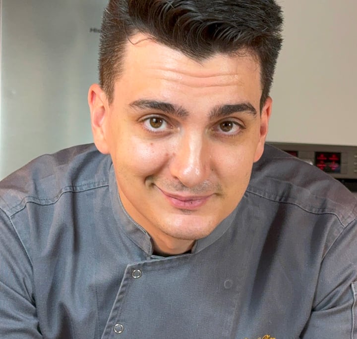 A person wearing a gray chef's jacket, standing in front of a modern kitchen setup with stainless steel appliances and an oven in the background.