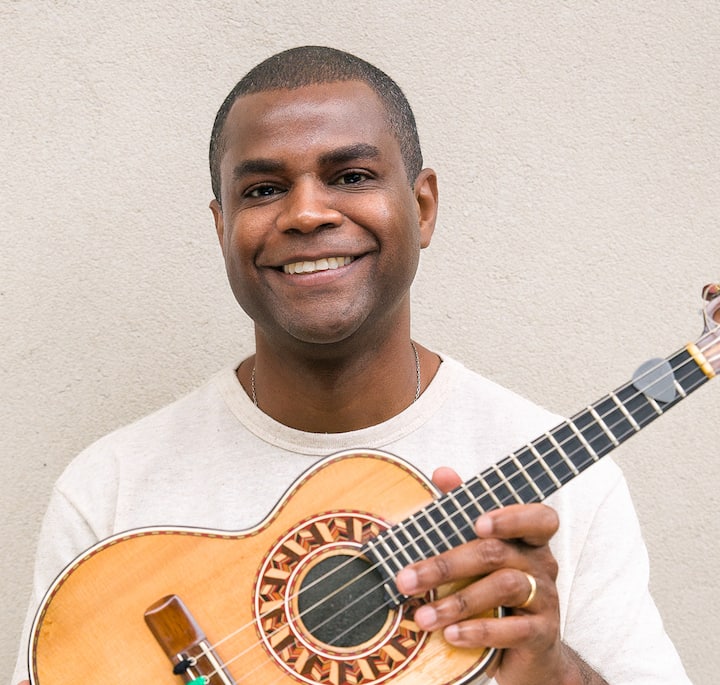 A close-up of a person holding a cavaquinho, a traditional Brazilian string instrument, with intricate wood detailing in the circular soundhole design, set against a neutral wall background.