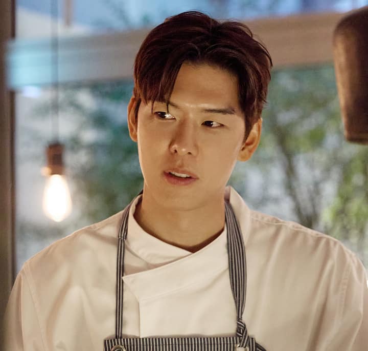 A chef wearing a white coat and striped apron stands under soft lighting, with blurred greenery visible outside the window behind him.