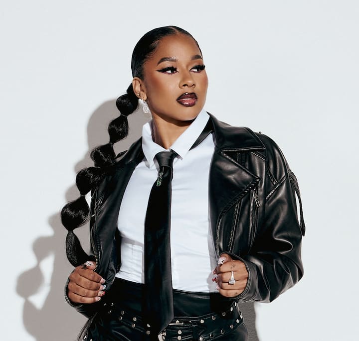 A person wearing a sleek black leather jacket over a white collared shirt and black tie, with a long braided hairstyle and studded black belt, posing confidently against a plain white background.