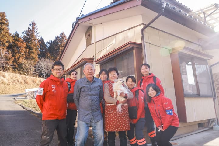 A group of disaster relief volunteers wearing red jackets stand with a smiling homeowner holding a small dog in front of a modest house surrounded by tall trees.
