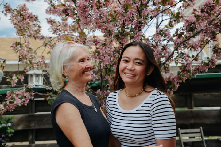 Two people standing outdoors in front of a blossoming cherry tree, surrounded by vibrant pink flowers and a wooden fence in the background.