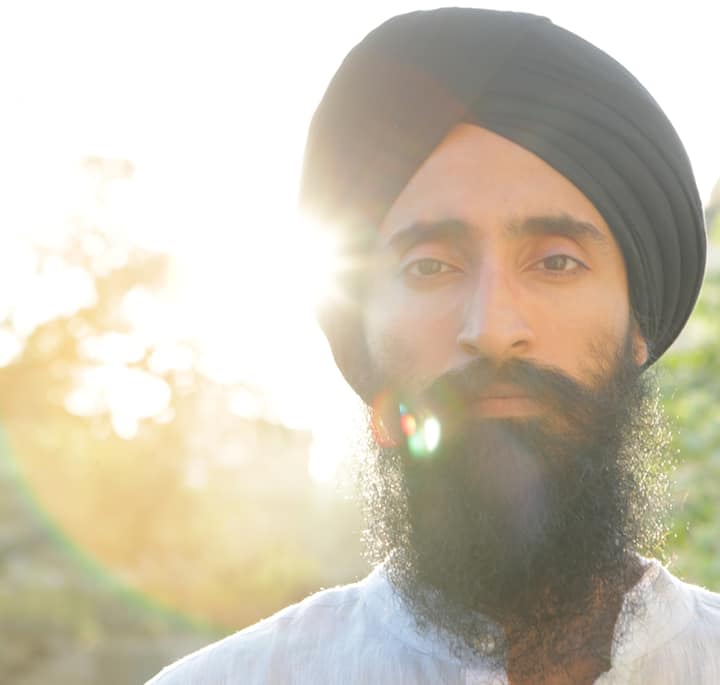 A person wearing a black turban and white clothing stands outdoors with sunlight shining through trees in the background.