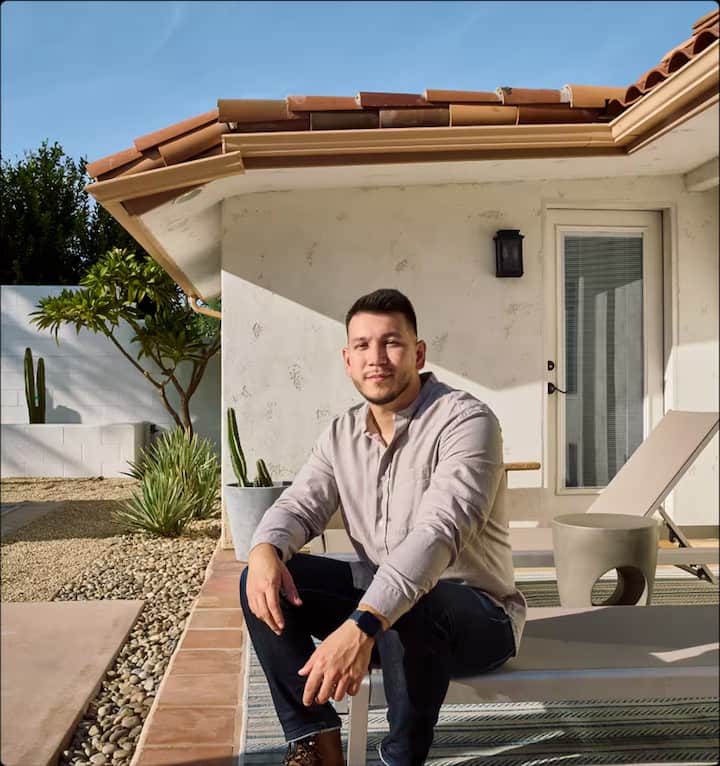 A man in dark pants and a tan top sits in a serene backyard with a lounge chair, potted plants, and a modern white stucco house featuring a terracotta-tiled roof and glass patio door, under a clear blue sky.
