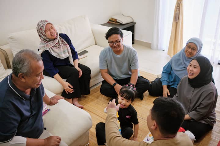 A warm gathering in a bright living room, with a group of adults sitting on a sofa and carpeted floor surrounding a young child who is playing with a toy. Sunlight streams through sheer curtains in the background.