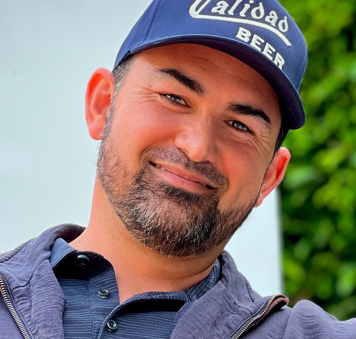 Close-up of a person wearing a navy blue cap with the embroidered text 'Calidad' and a casual dark jacket against a backdrop of vibrant green foliage.