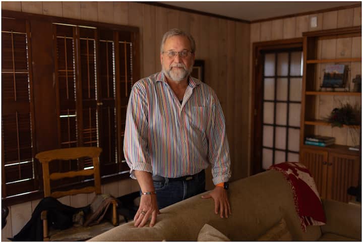 A man with a salt and pepper beard wearing a striped shirt stands in a cozy living room with wooden walls and furniture.