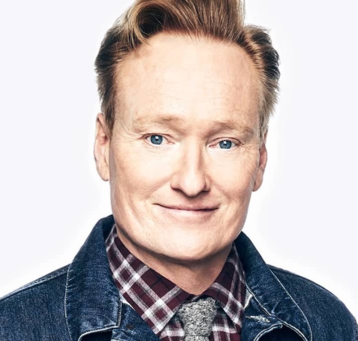 A close-up of a person wearing a denim jacket over a plaid shirt and tie, with the face blurred out, representing Conan O'Brien's Airbnb T1 experience.
