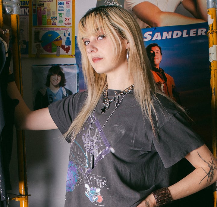 A person with long blonde hair wearing a vintage graphic t-shirt and layered necklaces, standing confidently in a room decorated with retro posters and colorful charts on the wall.