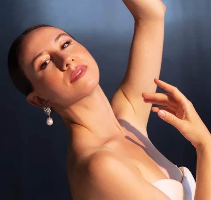 A close-up of a woman's arm and hand gracefully raised, wearing a strapless white outfit and pearl drop earrings. The lighting highlights her skin with a soft blue background.