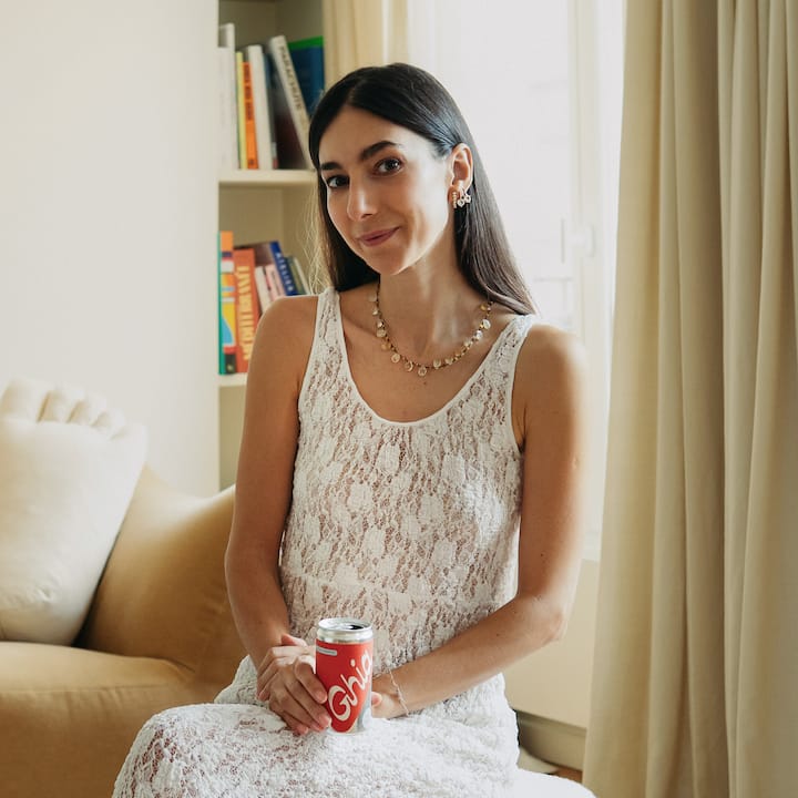 A woman wearing a white lace dress sits in a softly lit room holding a can of Ghia, with a beige sofa, bookshelf filled with colorful books, and sunlight filtering through curtains in the background.
