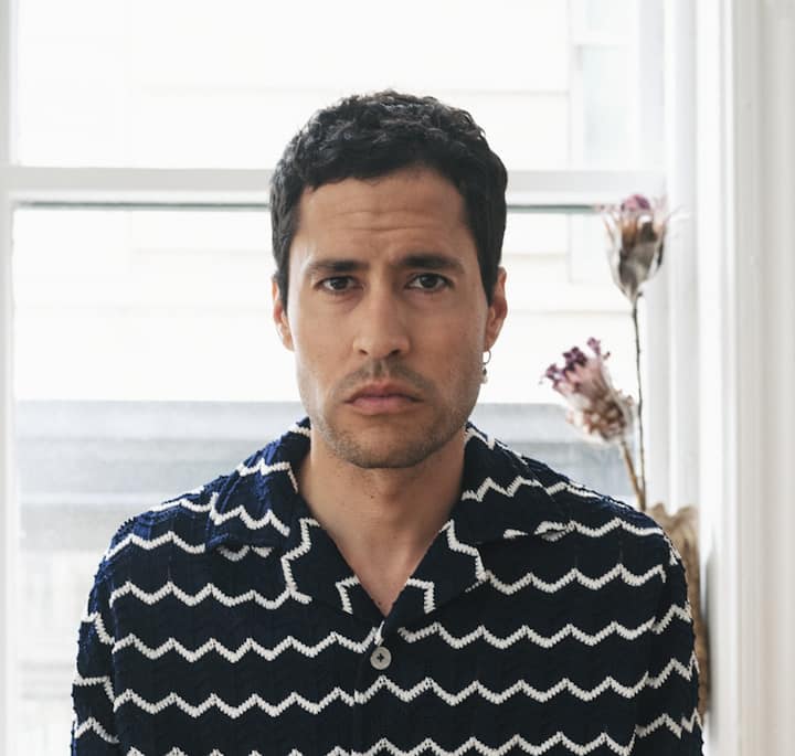 Portrait of an individual indoors, featuring a navy zigzag-patterned shirt, a white-framed window, and dried flowers in the background.