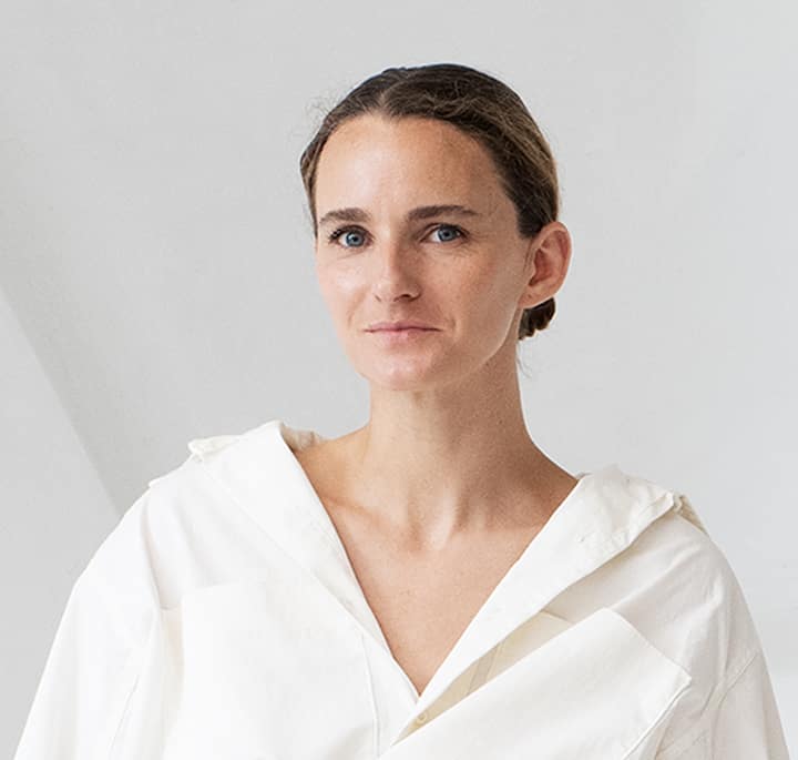 A close-up of someone wearing a minimalist, white, oversized shirt with a V-neckline and a relaxed collar, set against a neutral gray background.