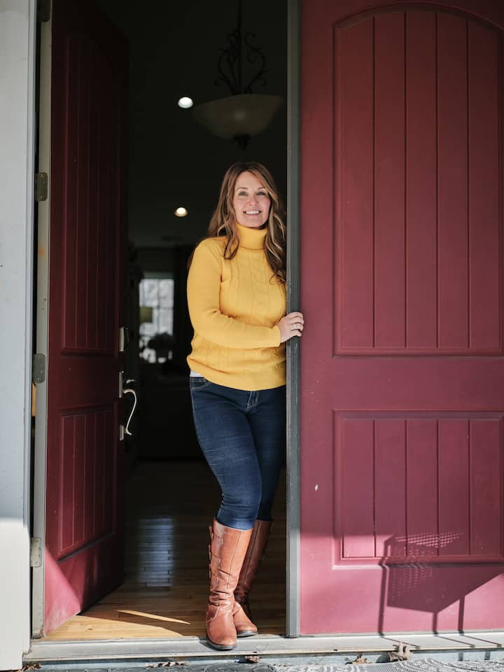 Airbnb.org host, Melissa, opened her home to volunteers during Hurricane Helene. She is standing in a yellow sweater next to a red door.