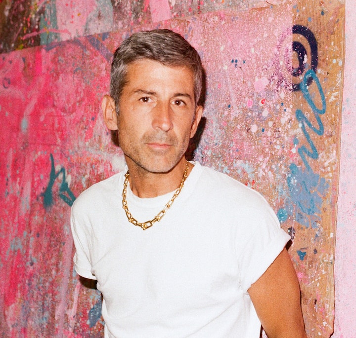 A man wearing a white t-shirt and gold chain stands in front of a graffiti-covered wall with pink, blue, and beige tones.