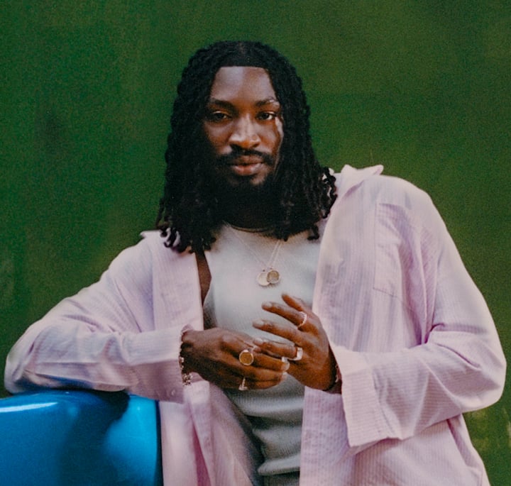 Person with long black locs wearing layered silver rings, a light pink shirt over a white tank top, and a silver necklace, leaning against a blue surface with a blurred green background.