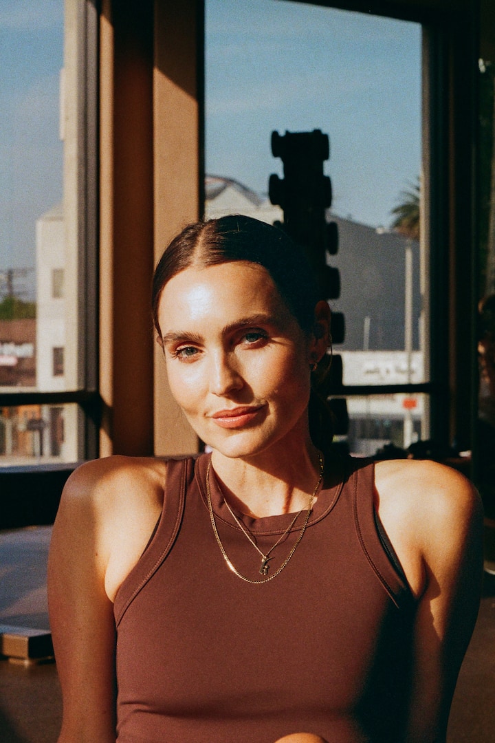 A person wearing a dark brown athletic tank top and layered gold necklaces sits indoors near a sunlit window, with urban buildings visible outside.
