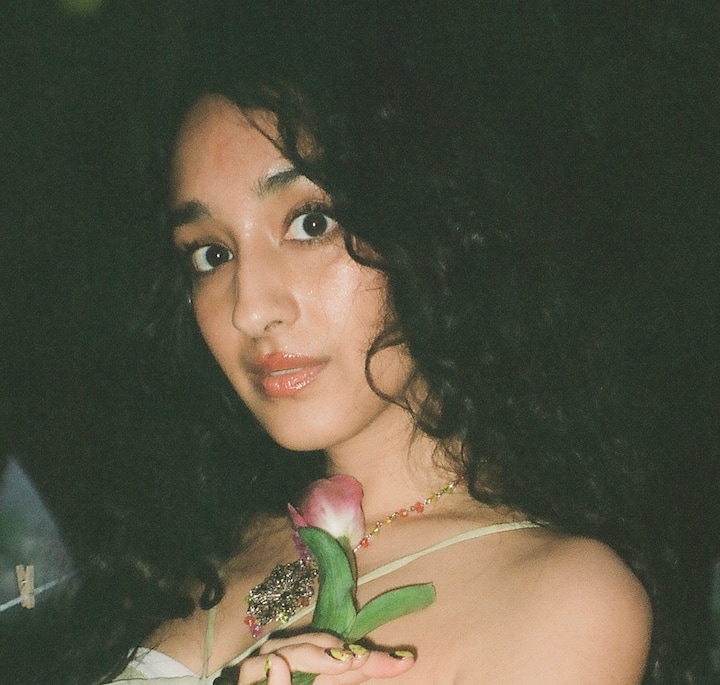 A close-up of a person with long curly hair holding a pink tulip flower, wearing a delicate necklace and a white dress, set against a dark background.