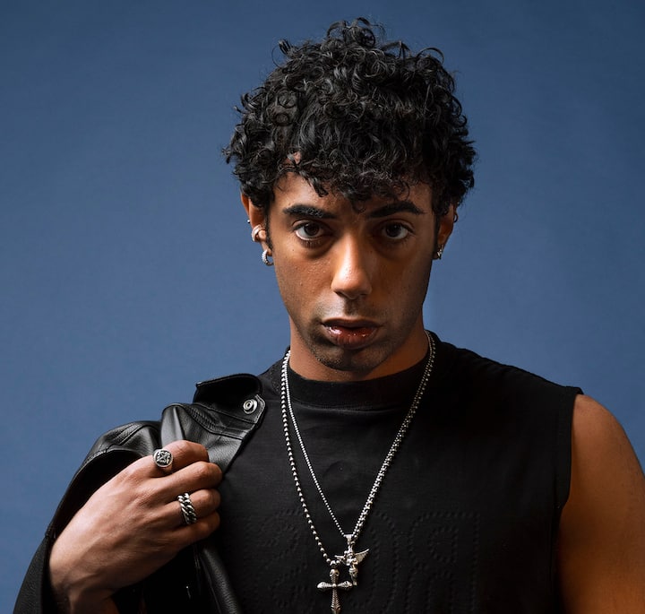 A person with curly black hair wearing a black sleeveless shirt, layered silver necklaces, and multiple rings, holding a leather jacket over their shoulder against a solid blue background.