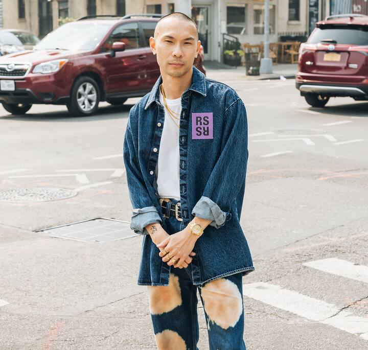 Person wearing a denim outfit with tie-dye-style pants and rolled-up sleeves standing on a city street, with cars and buildings in the background.