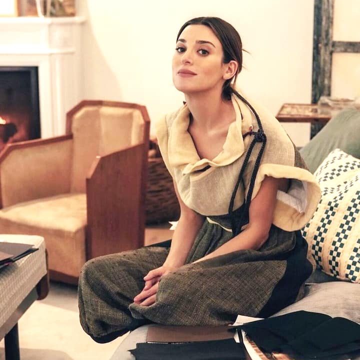 A stylishly dressed person sitting in a cozy room with a lit fireplace in the background, surrounded by neutral-toned furniture and fabric samples on the table, highlighting a fashion consultation setting.