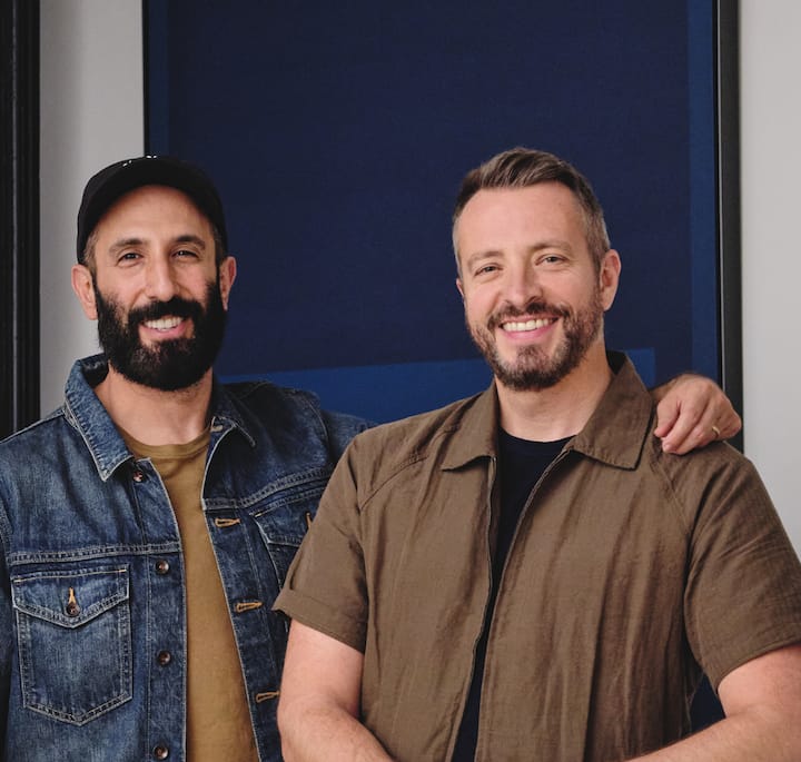 Two people standing side by side, one wearing a denim jacket and tan shirt, the other wearing a brown button-up shirt, with a dark blue wall in the background.