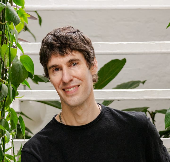 A person wearing a black shirt surrounded by green leafy plants in a minimalist setting with white stairs in the background.