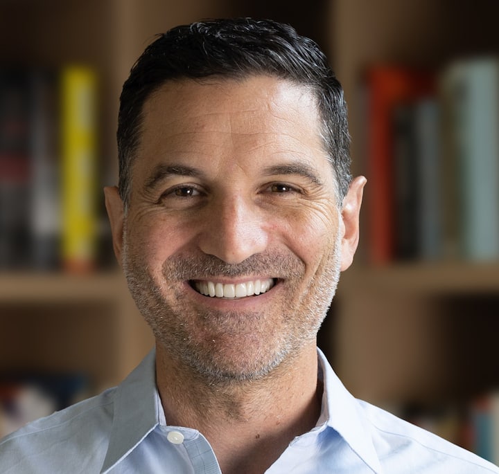 A close-up of a person wearing a light blue collared shirt, standing in front of a bookshelf filled with books of various colors.