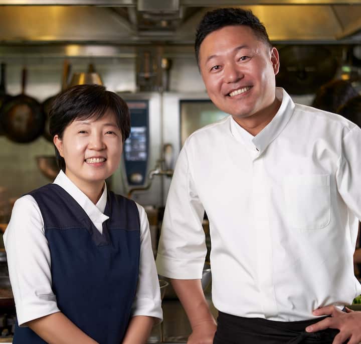 Two chefs in professional attire standing in a modern kitchen, with cooking equipment and utensils visible in the background.