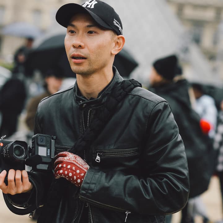 A person wearing a black leather jacket and red patterned gloves holds a professional camera in an outdoor setting with blurred people and an iconic glass pyramid in the background.