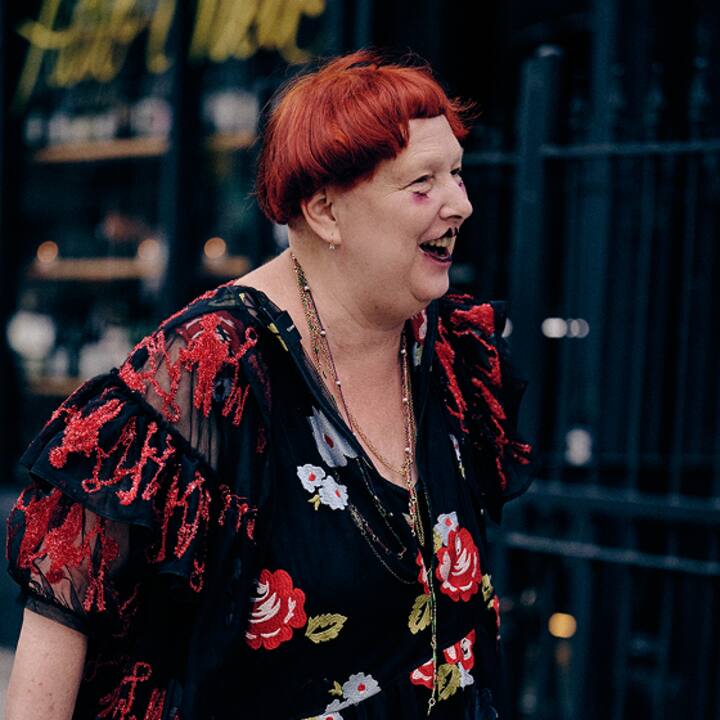 A person wearing a black dress with red and white floral patterns, standing outdoors near a building with a wrought iron fence in the background.