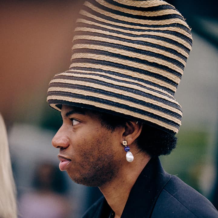 Close-up of a person wearing a black and beige striped hat and pearl earrings with a blurred outdoor background.