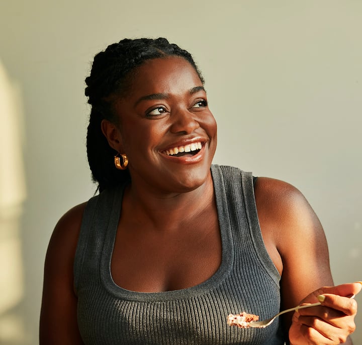 A person wearing a sleeveless gray top holds a spoon with a small serving of dessert, in soft natural lighting.