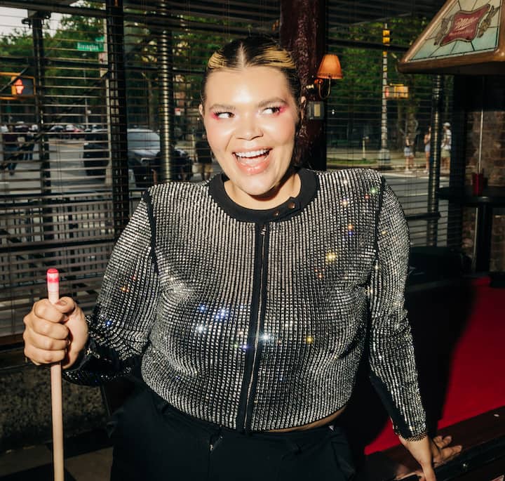 A person wearing a sparkling, metallic silver jacket holds a pool cue in a dimly lit billiards hall with large windows showing a city street outside.